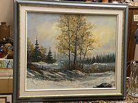 Framed vintage oil painting showing a winter landscape scene with trees and snow, resting on a table with a silver and gold frame.