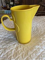 Front angled view of yellow ceramic pitcher showing handle and spout, placed on white quilted surface.