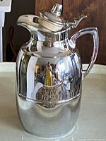 Full view of vintage thermal jug with reflective stainless steel body, ergonomic handle and lever lid.