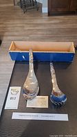 Two Acme Indian silver plated serving spoons on black surface with blue box behind. Includes ladle and fork spoon, tablet card description visible.