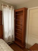 Tall pine wood bookcase placed beside a window with curtains and a door visible nearby, showing its full height and shelving layout.