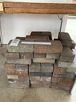 Stack of approximately 60 bricks showing faded and weathered surfaces in various brown and gray shades, arranged in a garage.