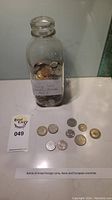 View of glass bottle filled with various foreign coins and some coins laid out in front of it. The label states the coins are a mixed collection from Asian and European countries.