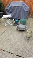 Overall lot showing white folding table, blue watering can, green bucket, black metal plant stand, shallow metal fire pit bowl. BBQ grill covered in background not included.