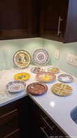 Ten decorative plates arranged on a white countertop in a kitchen corner. Plates vary in size and style with detailed handcrafted designs including floral, animal, geometric and cultural motifs. Two clear acrylic display stands are visible behind the plates.