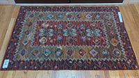 Full view of rectangular hand tufted wool area rug in muted reds, blues, yellows, and creams with floral and geometric design laid on hardwood floor.
