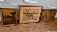 Three framed inlaid wood art pieces on floor; Old Mill scene on left, Loon in middle, Village scene on right.