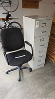 Photo of a black office chair positioned next to a tall beige metal 4-drawer file cabinet in a garage setting, showing chair's upholstery and cabinet drawers with handles and label holders.