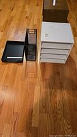 Three office organizers displayed on wooden floor including white multi-shelf unit, black mesh drawer organizer, and black desk tray