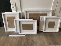 Seven white picture frames arranged standing and laying on wood floor, showcasing different rectangular and square sizes.