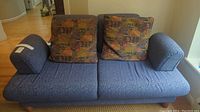 Blue ScanDesigns sofa with two multicolor patterned back cushions and padded blue armrests, shown against hardwood floor and beige walls