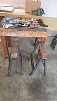 Assorted hand tools laid out on a table, with two metal sawhorses underneath. Shows a variety of hand tools and clamps.