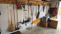 Wide view showing assorted yard tools including rakes, shovels, broom, wheelbarrow, oil containers, bins and hanging items in garage