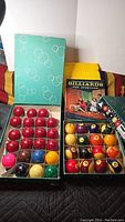 Two sets of billiard balls displayed side by side in their original boxes on a striped fabric.
