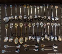 A full layout of 32 collector souvenir spoons arranged in rows on a flat surface with various crests and detailed bowls