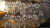 Full view of 44 collector spoons arranged on a surface showing various designs and emblems representing different locations globally. Spoons vary in size and decorative features.