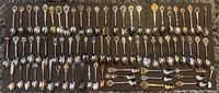 Overview of approximately 57 collector spoons laid out in rows on a dark surface, showcasing various handle designs and bowl shapes.