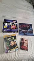 All four items neatly arranged together on a bed sheet: Mind Trap board game, Dinosaur Disney book, CSI DVD Game, Dexter Final Season DVD.
