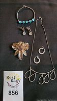 Photo showing all jewelry items: bracelet with turquoise beads, drop earrings, floral brooch with purple stones, silver hoop earrings, and silver necklace with teardrop-shaped pendants.