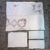 Photo of white and cream table linens including a large embroidered tablecloth with floral and lace crochet details, plus two smaller matching napkins with similar embroidery.