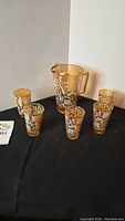 Full set of one amber-tinted glass pitcher with floral etchings and five matching glasses on a black fabric background.