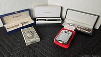 Overview of all items in the lot showing Waterman Paris pen in blue box, Balmain pen and pen set in white cases, and Restoration Hardware hand warmers with box and pouch.