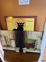 Framed print showing a black cat illustration with some French text, behind two canvas prints leaning on a table.