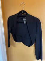 Black Frank Lyman bolero jacket hanging on a hanger, showing front with open cropped fit and long sleeves.