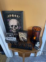 Photo showing Halloween decor arranged on a small wooden side table against a beige wall including framed skull art, wooden Halloween blocks, 'WICKED' sign, a skeleton dangler, raven block and a themed candle holder.