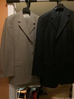Two men’s suits, beige and black blazers hanging side by side, each with matching pants.