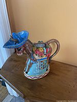 Side view of donkey shaped ceramic pitcher wearing vibrant blue sombrero, showing floral decoration and handle.