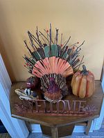 Thanksgiving decorative items arranged on a wooden small table or stand near a wall corner. Items include a fabric turkey centerpiece, ceramic pumpkin, 'WELCOME' sign, and raccoon figurine with natural elements.