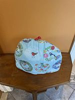 Tea cozy displayed on wooden table showing front with bird and wreath designs on light blue fabric with red loop on top.
