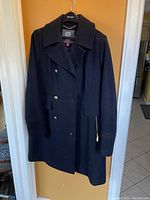 Full front view of navy blue Vince Camuto military style dress coat hanging on hanger on door. Shows buttons and slight texture.