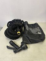 Photo showing the coiled black expandable garden hose, brass fitting, black spray nozzle with trigger, and black storage bag with clear window.