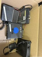 Photo of a box containing three CB radios including a Motorola unit with heat sink and coiled microphone cable, visibly used condition.