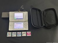 White Nintendo DS with dual screens powered on showing user name input screens, with AC charger and black carrying case next to it, five Nintendo DS game cartridges aligned in front.