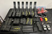 Top view of various slot car racing track pieces, two white slot cars, hand controllers, power adapter, and spares in a bag.