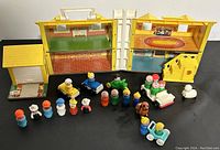 Two Fisher Price Little People fold-out house playsets with multiple vintage Little People figures including firefighters, clowns, adults, children, dog, sheep, and small cars in front of and around the playsets on a black surface.