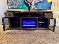 Front view of wooden entertainment center with flat screen TV placed above and LED heated fireplace insert glowing blue and purple in center. Drawers and cabinets open showing shelves inside.