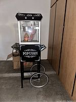 Front view of the complete popcorn machine showing black and silver carnival style design with wheels and handle.