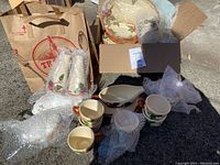 Photo of multiple Franciscan Apple patterned dish pieces, mostly wrapped in bubble wrap, placed on asphalt surface with a cardboard box and paper bag nearby.
