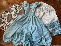 Three clothing items laid on wood floor: light blue dress with puff sleeves, white skirt with blue ruffles, and a floral quilt partially visible.