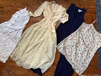 Four vintage women's dresses laid out: white lace slip, cream lace formal dress, navy blue sleeveless dress, white floral slip dress