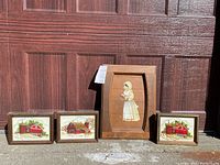 Four framed artworks displayed outdoors against wooden background. Three show red barn scenes in crewel embroidery style, one is folk art baker figure on wood.