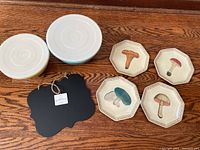 Set of four octagonal melamine plates with distinct mushroom illustrations, two melamine bowls with white lids, and one unused black scalloped chalkboard with hanging string
