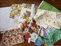 Photo showing assortment of mid-century linens in floral, autumn, and green color palettes, including napkins, placemats, and cloths spread out on wooden floor.