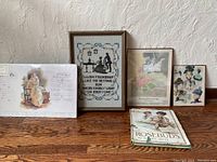 Five items arranged on wood surface: framed needlepoint with sentimental embroidery, framed prints including Victorian fashion hats (1898 Delineator), baby print, and books (Ladies Home Journal and Rosebud).