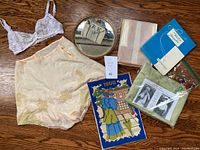 Overview photo showing bloomers, white lace bra, mirror with cross stitch purse, nylons box, scarf packaging, and blue tag with lot number