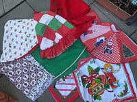 Photo showing an assortment of Christmas tree skirts and linens on a paved surface, including colorful tree skirts with patchwork and holiday prints, folded tablecloths, and several linen items.